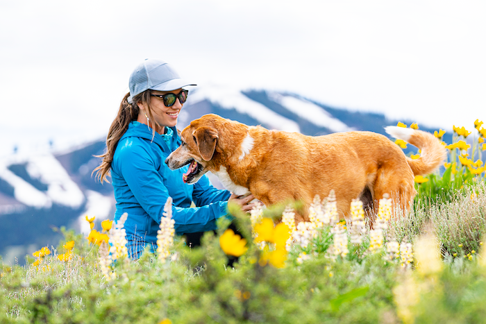 Woman crouches among with wildflowers with her dog with a snow-capped Bald Mountain in the background